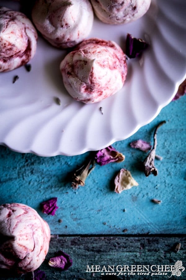 Overhead photo of Strawberry Meringue Cookies sitting on a white plate that's placed on old blue boards and sprinkled with flower petals.