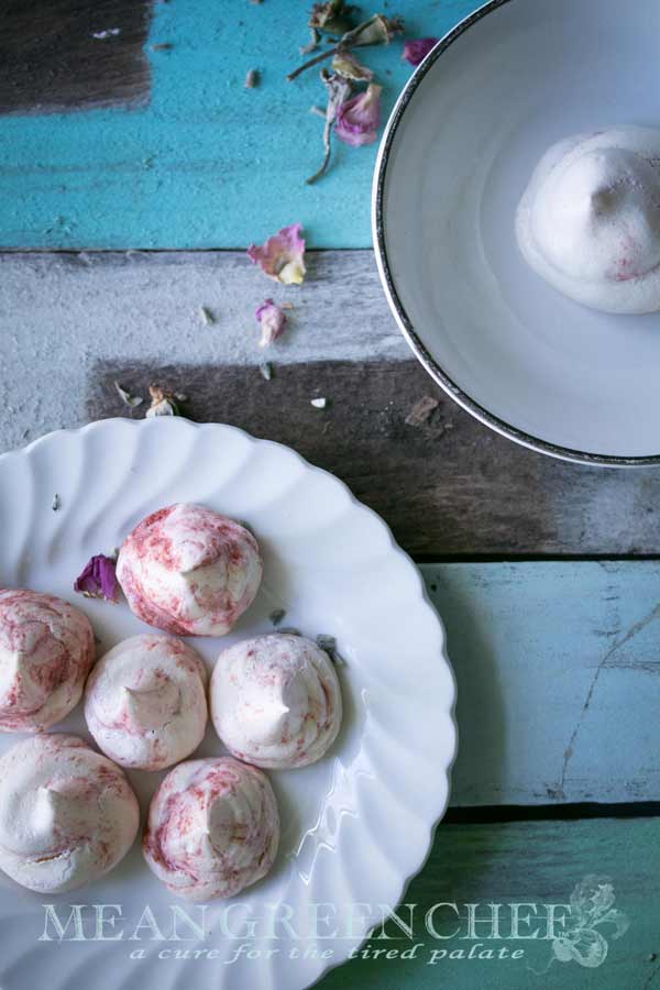 Overhead view of Strawberry Meringue Cookies with swirls of strawberry purée sitting on a scalloped white plate that's placed on rustic blue boards that has been sprinkled with small rose petals and lavender.
