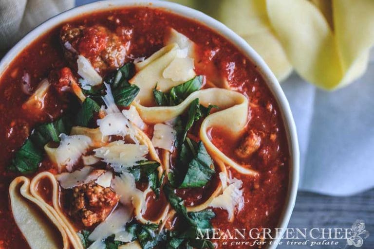 Overhead photo of Lasagna Soup garnished with fresh basil and homemade egg noodles with a scalloped spoon on a gray background.