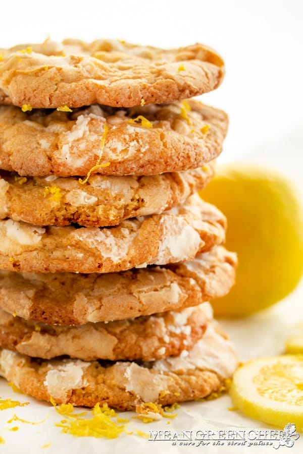 A stack of Meyer Lemon Crinkle Cookies with visible lemon zest and powdered sugar, set against a white background with slices of fresh Meyer Lemons at the base.
