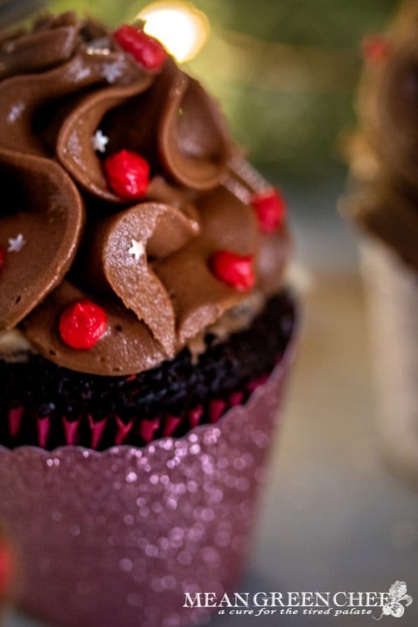 Side photo of Bakery Style Chocolate Cupcake decorated with a high swirl of chocolate frosting red frosting berries and a sprinkle of silver stars in a scalloped pink cupcake wrapper.