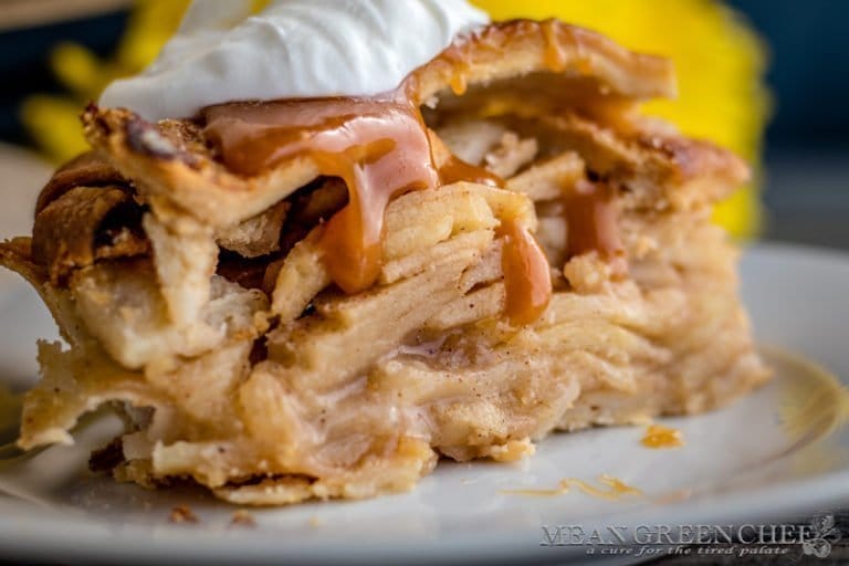 Side photo of a huge slice of Caramel Apple Pie on a white plate garnished with caramel and soft whipped cream. With a pie in the far background. Mean Green Chef