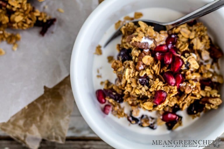 Overhead photo of a bowl of Brown Butter Granola in a white bowl garnished with pomegranate seeds on a wooden background. Mean Green Chef