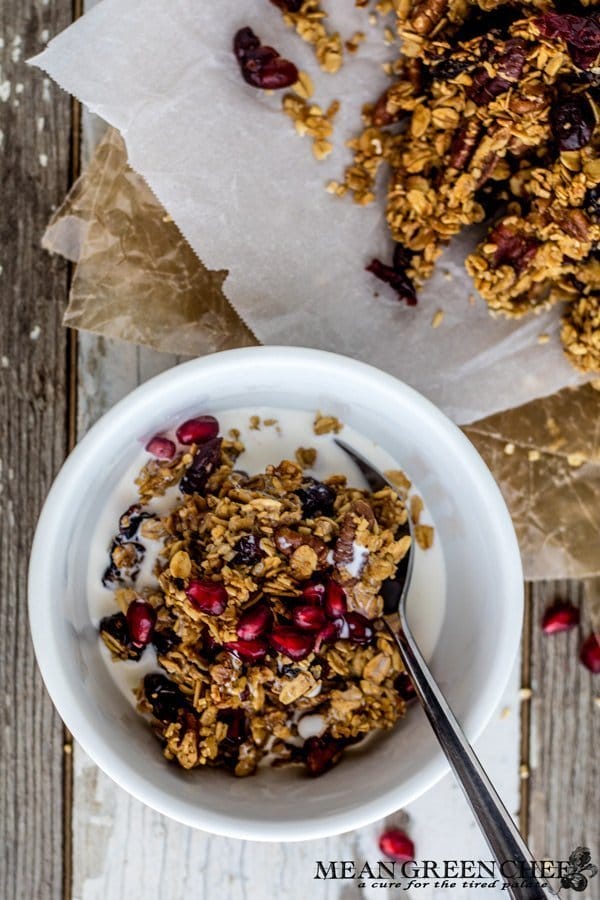 Overhead photo of a bowl of Brown Butter Granola in a white bowl garnished with pomegranate seeds on a wooden background. Mean Green Chef