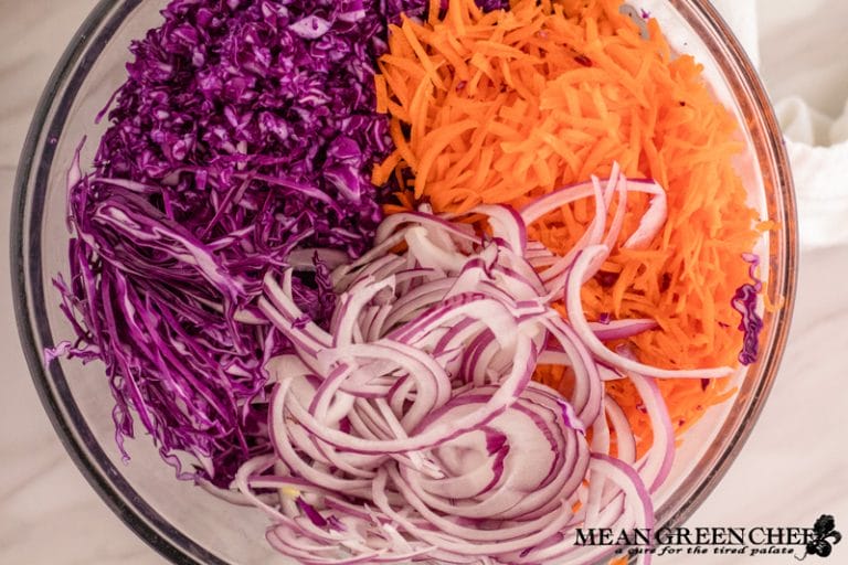 Ingredients for Red Cabbage and Carrot Slaw sliced in a large glass bowl on a white marble counter top background. Mean Green Chef
