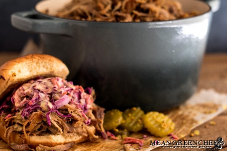 Side photo of Slow Roasted Pork Shoulder used on a pulled pork sandwich with red cabbage slaw and pickles off in the background. Mean Green Chef