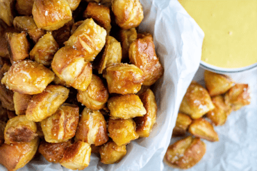 Close-up of golden brown pretzel bites in a white paper-lined bowl, accompanied by a cup of honey mustard sauce on the side, on a light textured surface.