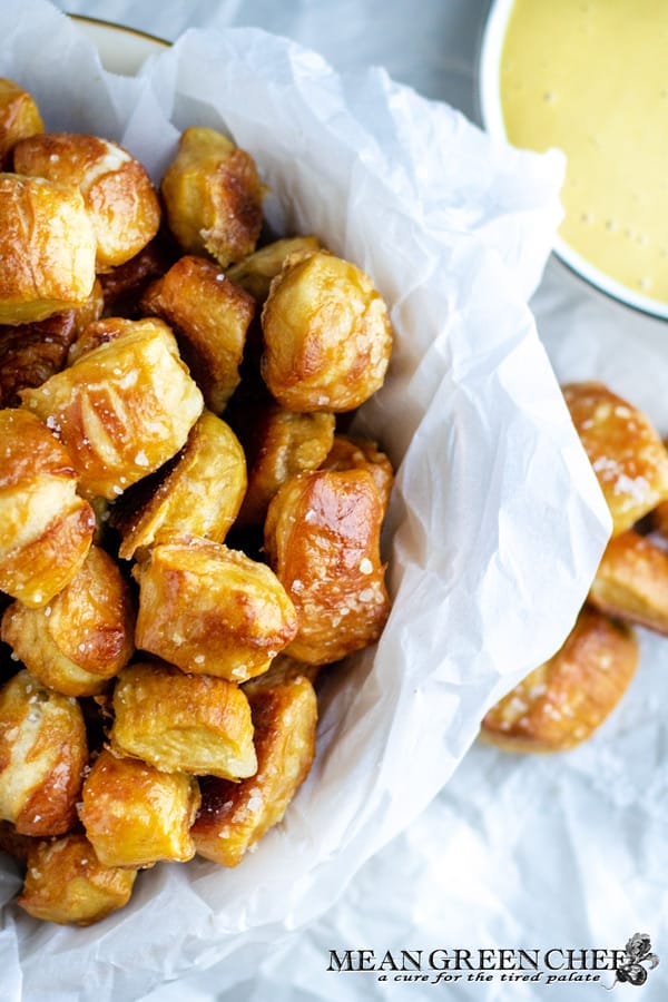 Close-up of golden brown pretzel bites in a white paper-lined bowl, accompanied by a cup of honey mustard sauce on the side, on a light textured surface.