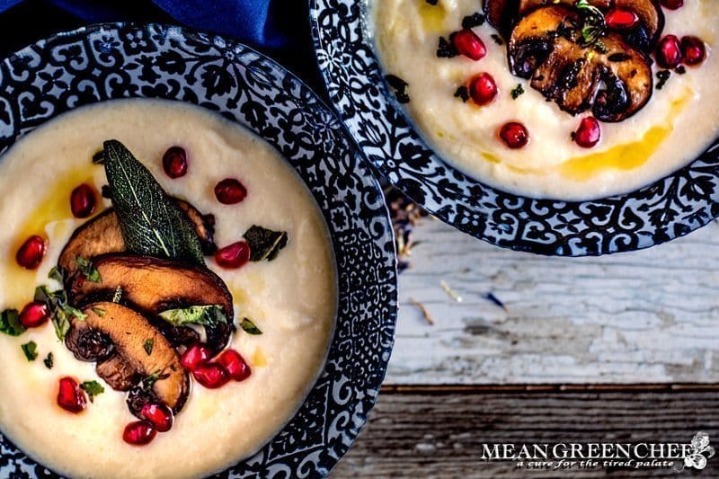 Creamy Cauliflower Bisque ladled into two blue bowls and garnished with smoked portobello mushrooms, fried sage, and pomegranate seeds.