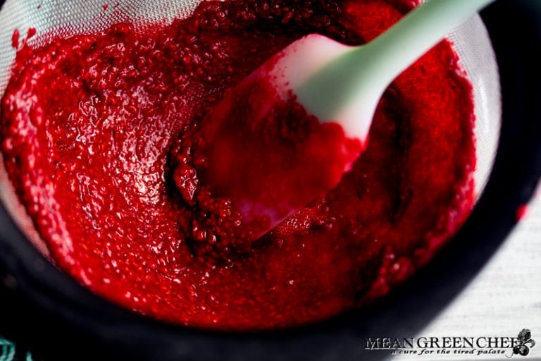 Overhead photo of raspberries being pushed through a fine mesh strainer into a glass measuring cup.