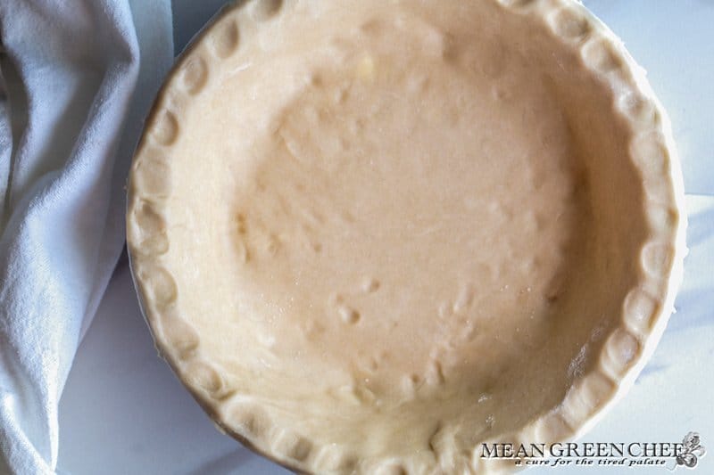 Bottom of a pie crust formed into a 9 inch pie dish, photographed on a parchment paper background with a white kitchen linen running up the left side. Mean Green Chef