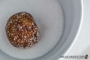 Close up of Best Ever Molasses Cookie dough rolled into a ball and coated in superfine white sugar in a white bowl on a white marbled background countertop.