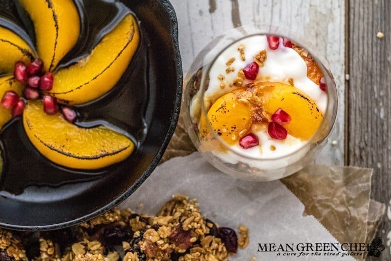Overhead photo of Easy Peach Parfait with Brown Butter Granola, Roasted Pecans, and Cranberries in a short, stout glass. With a small cast iron pan int he upper left hand corner showing off browned peaches. Mean Green Chef