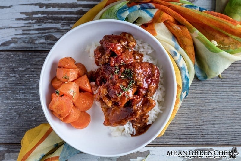 A bowl of Chicken Pierre coated in a rich tomato sauce, served over Jasmine rice. Garnished with fresh thyme, accompanied by a side of roasted carrots. Situated on a rustic wood background, with colorful silk woven underneath the bowl and out of the top of the frame.