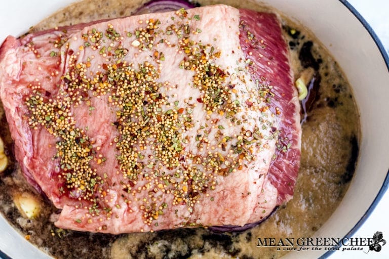 Raw corned beef roast seasoned with a mix of herbs, sitting on top of garlic cloves and bathed in Guinness Draught Stout. In a large blue-gray Dutch Oven.