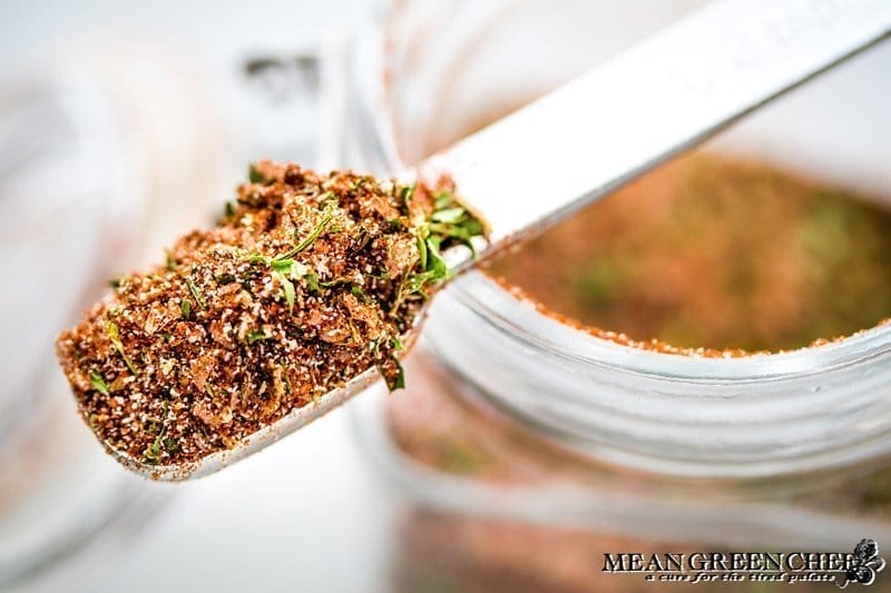 Close-up of a spoonful of mixed spices held above a glass jar, showing a detailed texture of Badass Blackened Seasoning with visible herbs and spices.