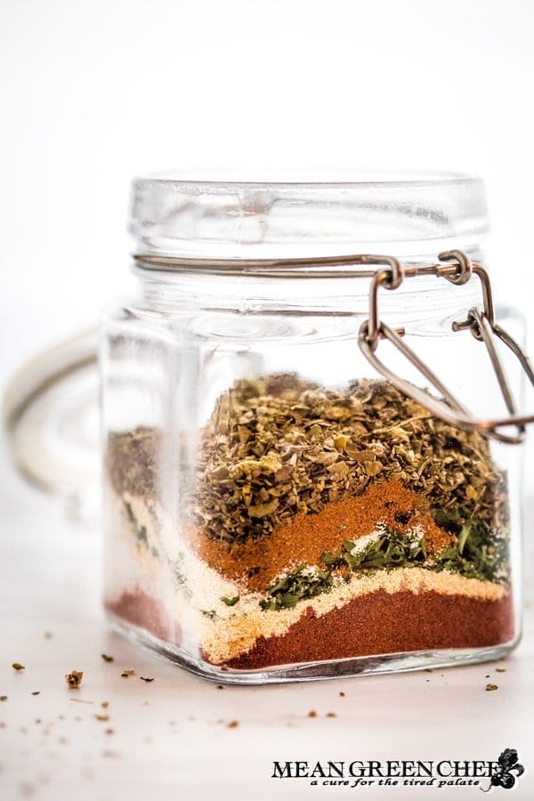 A glass jar with a clasp lid containing layers of colorful spices for making Badass Blackened Seasoning, including red, orange, and green, against a light, minimalist background.