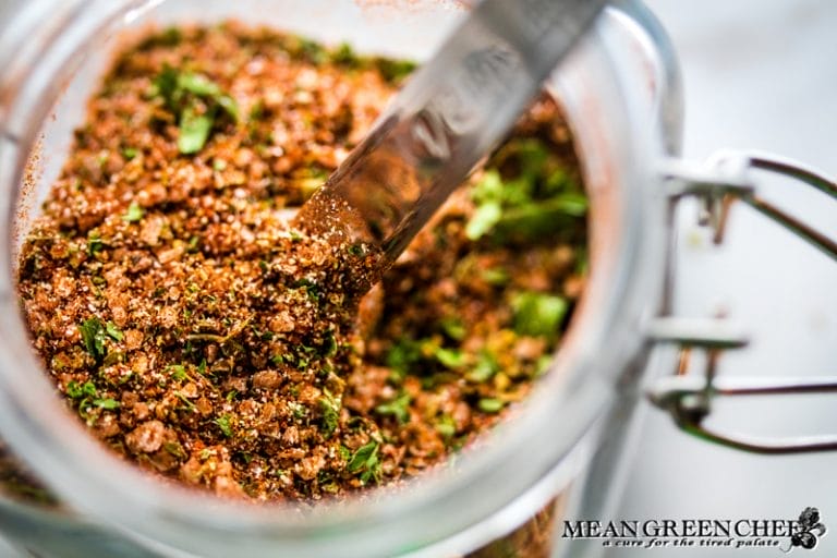 A close-up view of Badass Blackened Seasoning spice blend in a glass jar with a spoon, featuring visible herbs and spices, with the jar slightly open on a marble countertop.