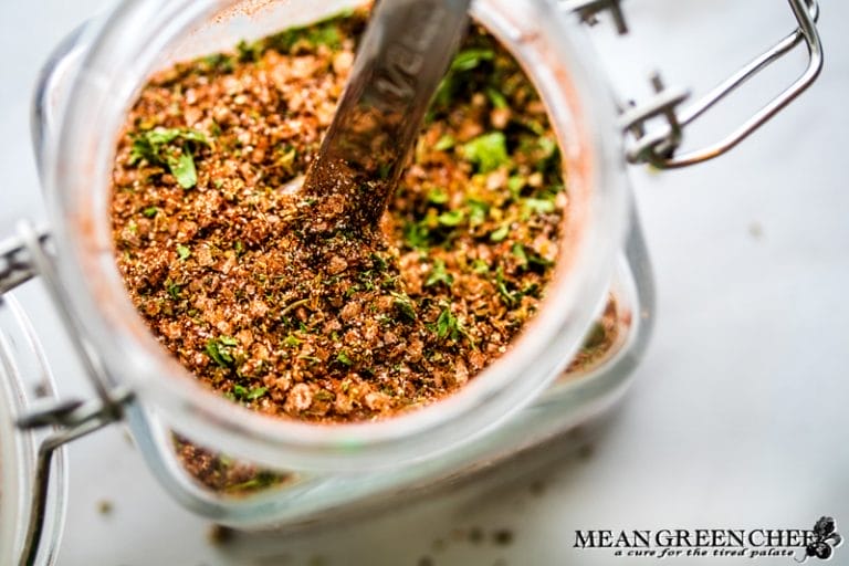 A close-up view of Badass Blackened Seasoning spice blend in a glass jar with a spoon, featuring visible herbs and spices, with the jar slightly open on a marble countertop.
