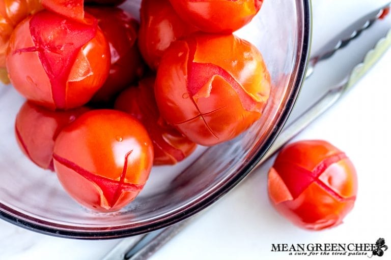 A large glass bowl of bright red blanched tomatoes with skins partially peeled, resting on a white surface next to a metal pair of tongs.