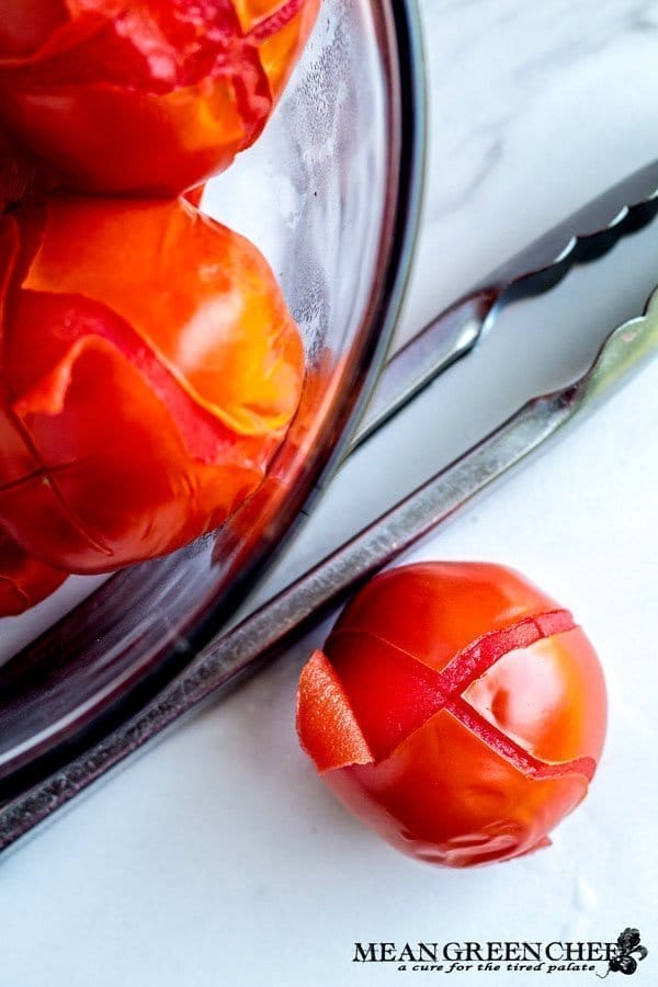 A close-up image of bright red blanched tomatoes, several in a glass bowl and another beside it, on a white marble countertop with a pair of silver tongs laying alongside the bowl.