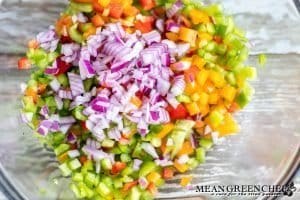 Green, red, and yellow bell peppers being tossed with a diced red onion for chicken pasta salad.
