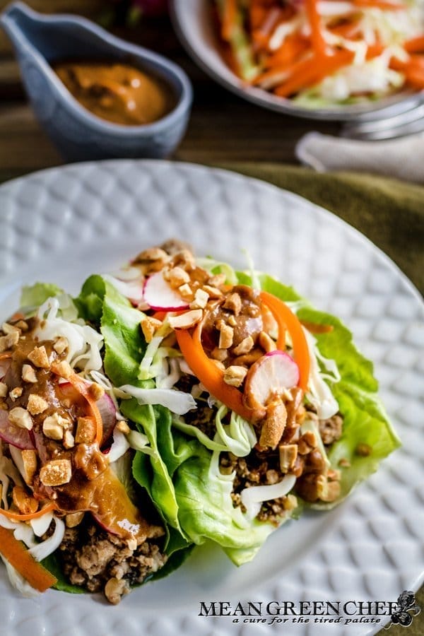 A vibrant dish of Thai Lettuce Wraps filled with meat, topped with spiralized carrots, radishes, nuts, and a drizzle of peanut sauce, served on a white plate with condiments on the side.