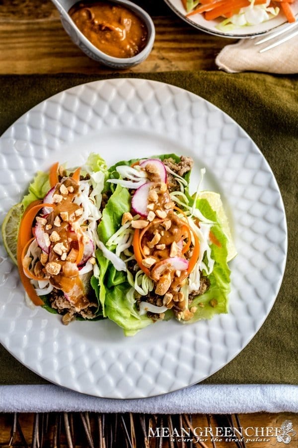 A vibrant dish of Thai Lettuce Wraps filled with meat, topped with spiralized carrots, radishes, nuts, and a drizzle of peanut sauce, served on a white plate with condiments on the side.