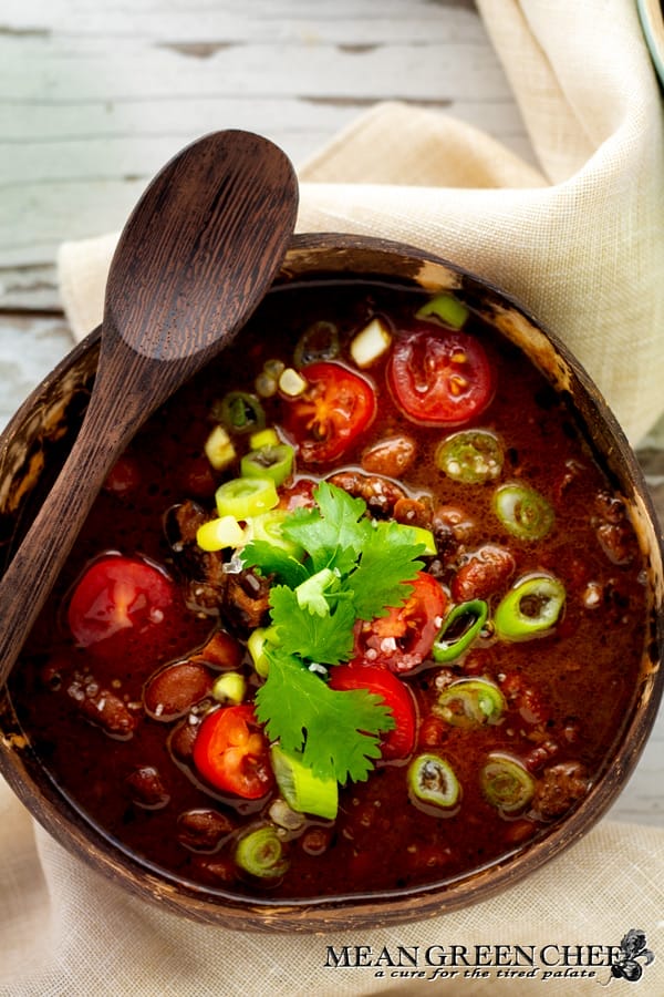 A bowl of Slow Cooker Texas Pinto Beans garnished with sliced jalapeños, cherry tomatoes, and fresh cilantro, accompanied by a wooden spoon, set on a rustic wooden table.