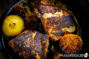 Closeup of pan-seared Gulfside Blackened Grouper fillets with a crispy blackened crust in a cast iron skillet, with a grilled lemon halves on the left-hand side.