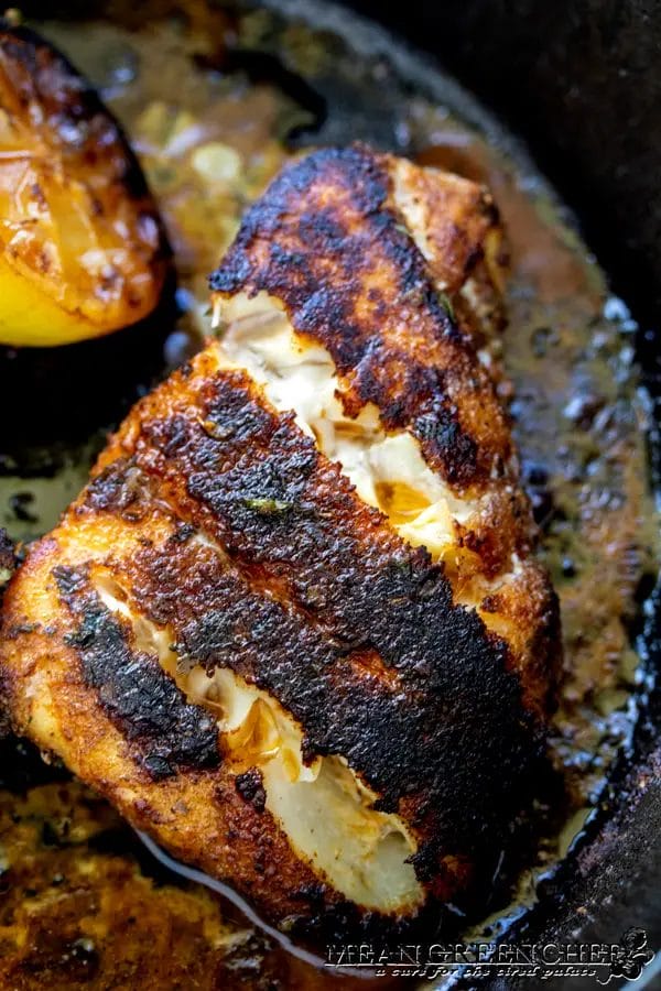 Closeup of pan-seared Gulfside Blackened Grouper with a crispy blackened crust in a cast iron skillet, with a half grilled lemon on the left-hand side.